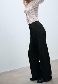 Femme portant un haut blanc en dentelle à manches longues, un pantalon noir à jambes larges et des mocassins noirs, debout sur un sol uni contre un mur blanc.