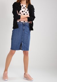 Jeanskjol i pennmodell med en framdelad split, kombinerad med en svart texturerad jacka och en rosa prickig skjorta, avslutad med rosa sneakers.