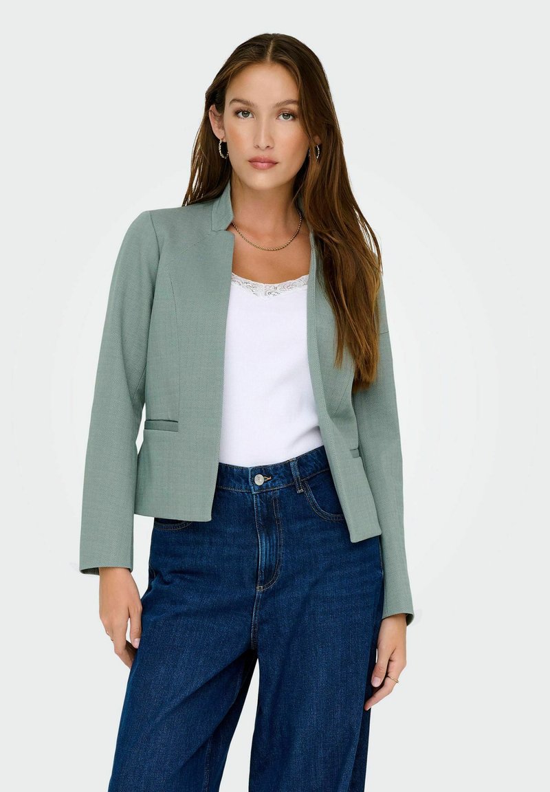 Femme aux longs cheveux bruns portant un blazer vert menthe, un haut blanc à bordure en dentelle, un jean taille haute bleu et des boucles d'oreilles créoles sur fond blanc.