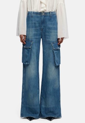 Blauwe wijde denim cargobroek met grote klepzakken op de dijen, gedragen met een witte blouse met lange mouwen en zwarte puntige schoenen.