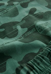 Tissu à motif camouflage vert présentant différentes nuances de vert, des points et des accents de texte. Le matériau semble doux avec un léger éclat.