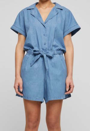 Femme portant une combinaison short en denim bleu clair à manches courtes, avec col et ceinture nouée à la taille, debout devant un fond uni.