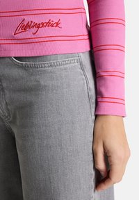 Rosa gerippte Langarmoberteil mit roten Streifen, versehen mit gesticktem Logo. Kombiniert mit grauen Denim-Jeans, die einen strukturierten Stoff zeigen.
