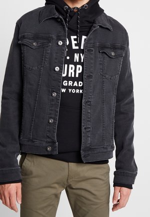 Mann trägt schwarze Jeansjacke über schwarzem Hoodie mit weißem Text, kombiniert mit beigefarbenen Hosen, stehend vor einem einfarbigen Hintergrund.