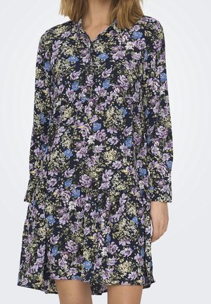 Frau trägt ein langärmeliges, knöpfbares Kleid mit Blumenmuster in Lila, Blau und Gelb auf schwarzem Hintergrund.