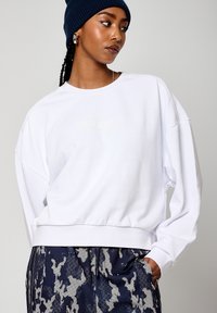 Felpa bianca con orlo a coste e spalle scese, caratterizzata da un logo sottile in colore chiaro. Abbinata a pantaloni navy a fantasia.