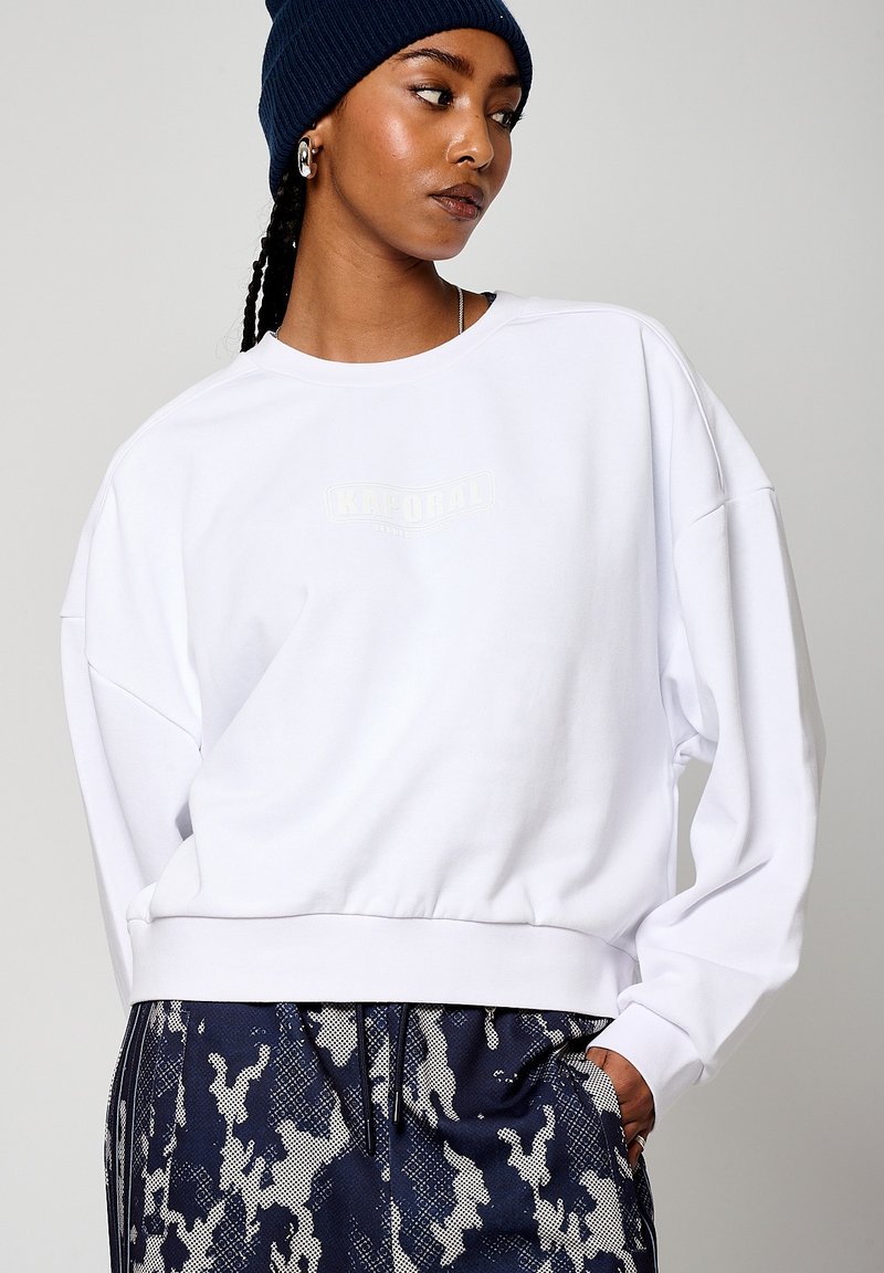 Felpa bianca con orlo a coste e spalle scese, caratterizzata da un logo sottile in colore chiaro. Abbinata a pantaloni navy a fantasia.