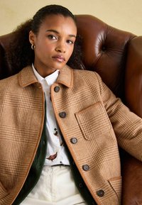 Brown houndstooth jacket with a structured collar, brown buttons, and a chest pocket, layered over a patterned shirt and green sweater.