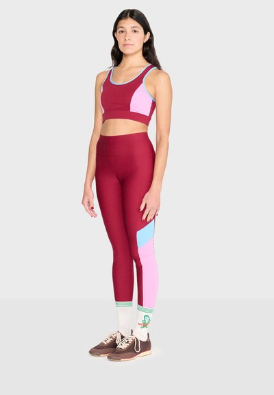 Femme debout portant un soutien-gorge de sport bordeaux, rose et bleu ainsi que des leggings, des chaussettes blanches à rayures vertes et des baskets marron.