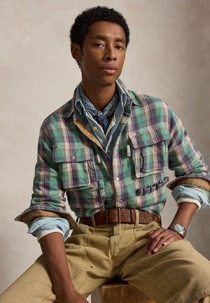 Jeune homme portant une chemise à carreaux verte par-dessus une chemise en jean aux manches retroussées, un foulard bleu, un pantalon beige et une montre, assis sur un tabouret.