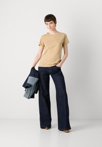 Beige katoenen T-shirt met korte mouwen en subtiele logo, gecombineerd met donkerblauwe flare jeans. Blauwe spijkerjack over één arm gehangen.