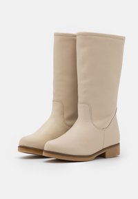 Botas de cuero beige hasta la rodilla con una textura suave, punta redondeada y un tacón de madera bajo. Presentan una costura mínima y un diseño clásico.