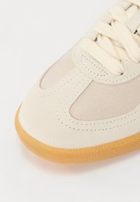 Beżowe sneakersy z zamszowymi i siateczkowymi panelami, kremowymi sznurowadłami oraz gumową podeszwą z teksturowanym wzorem.