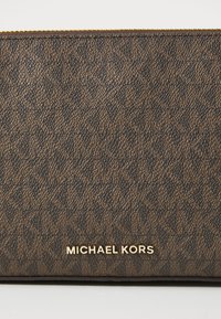 Brązowa, teksturowana skórzana saszetka z zamkiem, zdobiona wzorem z przeplatającymi się logo oraz złotą tabliczką z napisem "Michael Kors" z przodu.
