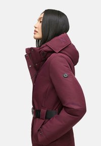 Bordeaux winterjack met een capuchon, ceintuurborst, ritszakken en logo-accenten. Kenmerkt zich door een gladde, gestructureerde stof en drukknoopsluitingen.