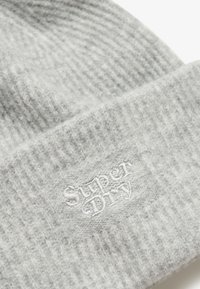 Gorro de punto gris con textura acanalada y un pequeño parche bordado que muestra "Super Dry". Material suave, vista en primer plano.
