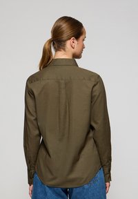 Olivgrön långärmad skjorta med klassisk krage, bakre yoke och knappade cuffs, med en slät textur och en något böjd nederkant.