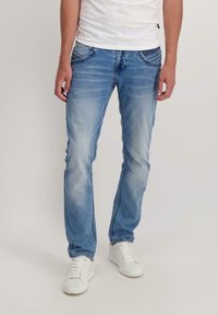 Man draagt lichtblauwe verweerde jeans, witte sneakers en een wit T-shirt, staand tegen een vlakke lichte achtergrond.
