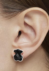 Orecchini a forma di orso nero con design sfaccettato e contorno argentato, posizionati contro un primo piano di un orecchio.