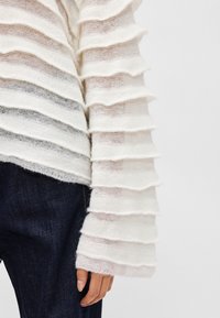 Pull en tricot blanc texturé avec un design à volants superposés. Matière douce avec une coupe ample, associé à un denim foncé pour le contraste.