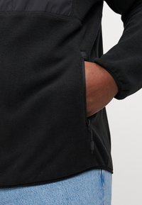 Black fleece jacket with a smooth texture, featuring a zippered side pocket and a contrasting black panel at the upper front.