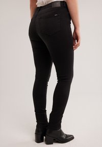 Cilvēks valkā melnus šaurus džinsus un melnas augstpapēžu potīšu zābakus, stāvot nelielā pagriezienā, rādot muguras un sānu skatu.