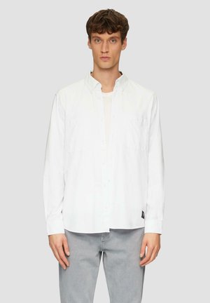 Chemise blanche à manches longues, avec deux poches poitrine et un ourlet arrondi. Portée sur un t-shirt de couleur claire. Tissu doux et lisse.