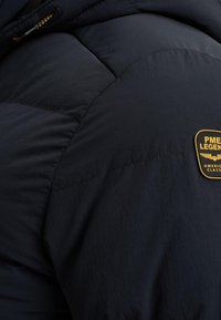 Zbliżenie na ciemnoniebieską kurtkę puchową, z pikowanym wzorem, widocznymi szwami oraz złotym logotypem na ramieniu.