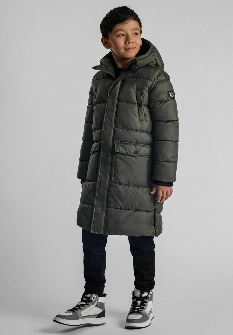 Zelena podložena bunda s kapuco, z vodoravno ožičenjem, dvema sprednjima žepoma in logotipom na rokavu. Nosi se z črnimi hlačami in športnimi copati.