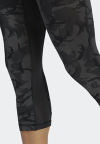 Zbliżenie osoby noszącej czarne i ciemnoszare legginsy sportowe w wzór kamuflażu z siateczkowymi panelami po bokach na jednolitym tle.