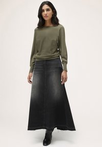 o bluză lungă cu mânecă, de culoare verde măsliniu, asortată cu o fustă maxi din denim negru, cu un efect de degradare și margine franjurată, purtată cu botine negre până la gleznă.