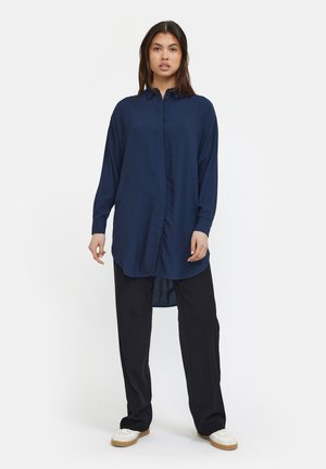 Navy blue long-sleeve button-up shirt with a curved hem, paired with black wide-leg trousers and white sneakers. Smooth fabric texture visible.