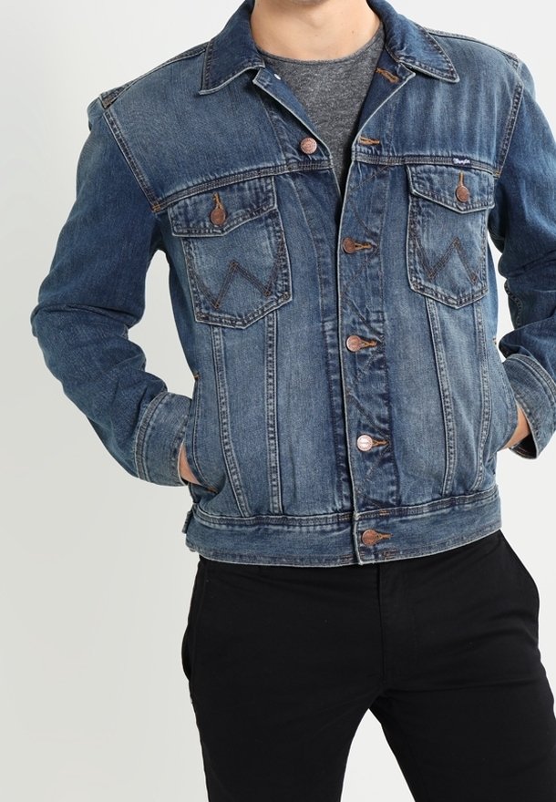 Mand iført en blå denimjakke over en grå skjorte med hænderne i jakkelommerne foran en ensartet lys baggrund.