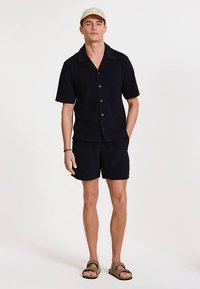 Camicia a maniche corte blu navy con bottoni, abbinata a pantaloncini coordinati. Presenta un tessuto strutturato e una vestibilità comoda. Indossata con un cappello chiaro e sandali.