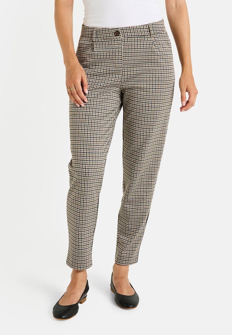 Femme portant un pantalon ajusté à carreaux beige et noir et des chaussures à talons bas noires, se tenant devant un fond blanc.