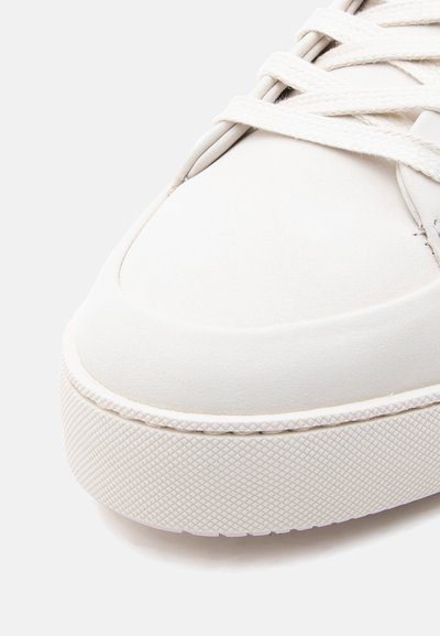 Λευκό δερμάτινο αθλητικό παπούτσι με υφή από καουτσούκ στ η σόλα, στρογγυλεμένη μύτη και επίπεδα κορδόνια. Ομαλή επιφάνεια με ελάχιστες ραφές και διακοσμητικά στοιχεία.