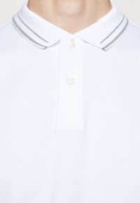 Polo blanc avec deux boutons et un col, ornée de deux fines rayures grises, portée par une personne visible du cou à la poitrine.