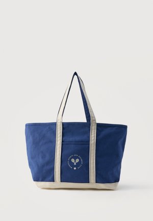 Sininen kassi, jossa on beigejä kahvoja ja pohja, etutasku sekä pyöreä logo, jossa on ristikkäiset tennismailat ja teksti.