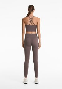 Bralette marrone sportivo con spalline incrociate e leggings coordinati a vita alta. Texture liscia, design aderente e accenti di logo sottili.