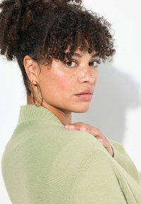 Ljusgrön stickad tröja med ribbad krage, med en närbild av en person med lockigt hår och stora hoopsmycken.