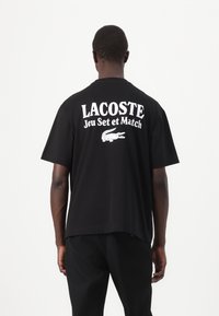 Svart bomull T-skjorte med "LACOSTE Jeu Set et Match" i hvit tekst på ryggen, med en logo av en krokodille nedenfor. Korte ermer, løs passform.
