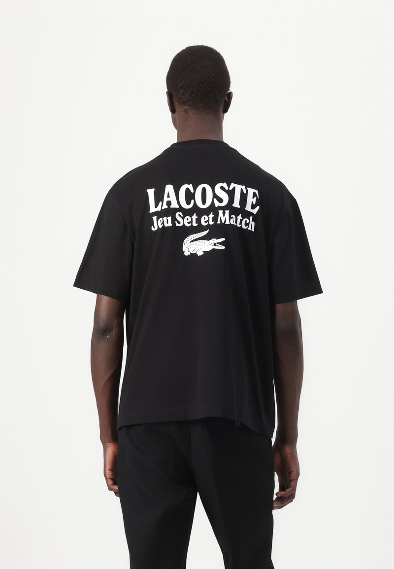 Svart bomull T-skjorte med "LACOSTE Jeu Set et Match" i hvit tekst på ryggen, med en logo av en krokodille nedenfor. Korte ermer, løs passform.