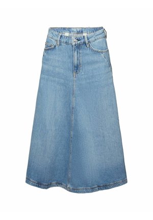 Femme portant un haut orange sans manches, une jupe midi en jean bleu clair et des sandales noires ouvertes à l'avant, debout devant un fond blanc.