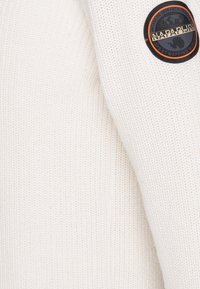 Valkoinen neulottu villapaita, jossa on teksturoitu kangas. Hiha on koristeltu pyöreällä mustalla läpällä, jonka reunassa on oranssi reunus ja ympyrämäinen kuvio.