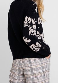 Marinblå stickad tröja med krämfärgade blommönster, rund halsringning och ribbad fåll. Bärs över rutiga byxor med flera färger och texturer.