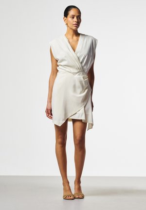 Frau trägt ärmelloses weißes Wickelkleid mit asymmetrischem Saum und braunen offenen Schuhen, steht vor einfachem hellem Hintergrund.