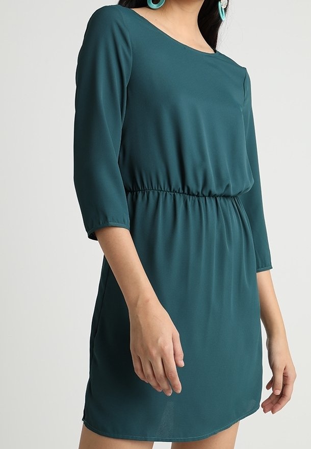 Femme portant une robe vert sarcelle à manches longues, longueur genou, avec une taille élastique, debout devant un fond clair uni.