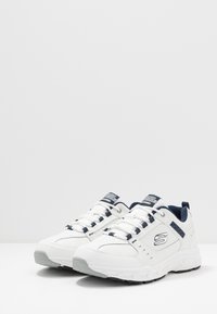 Scarpe da ginnastica bianche con accenti blu navy, materiale sintetico testurizzato, punta tonda e suola imbottita. Presentano lacci e dettaglio del logo sul lato.