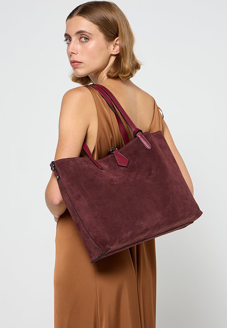 Burgunderfarbene Wildleder-Tote mit doppelten Griffen, glatter Textur, minimaler Hardware und strukturierter Form. Verfügt über einen Reißverschlussverschluss oben.