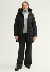 Manteau en duvet noir avec capuche, présentant un design matelassé et une fermeture éclair à l'avant. Porté sur un col roulé gris clair, associé à un pantalon large sombre.